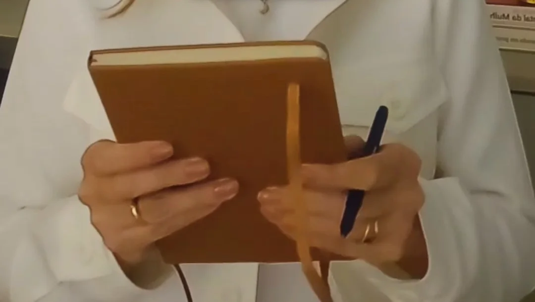 As mãos da Dra. Ana Emília segurando um caderno de capa marrom e uma caneta