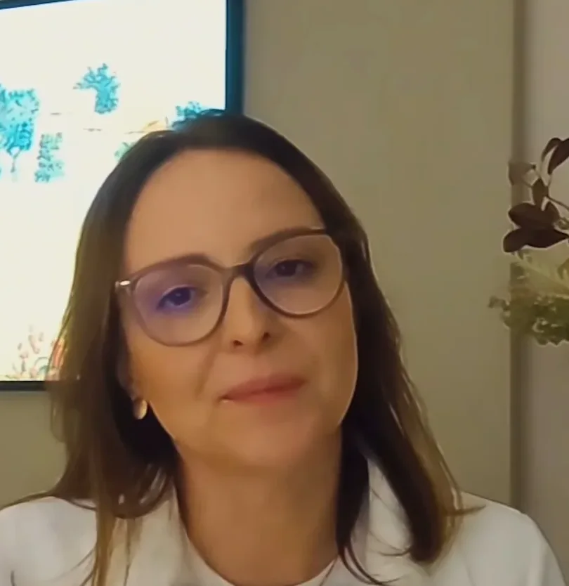 Dra. Ana Emília Pinheiro, de óculos e blazer branco, numa sala com um quadro de flores azuis ao fundo