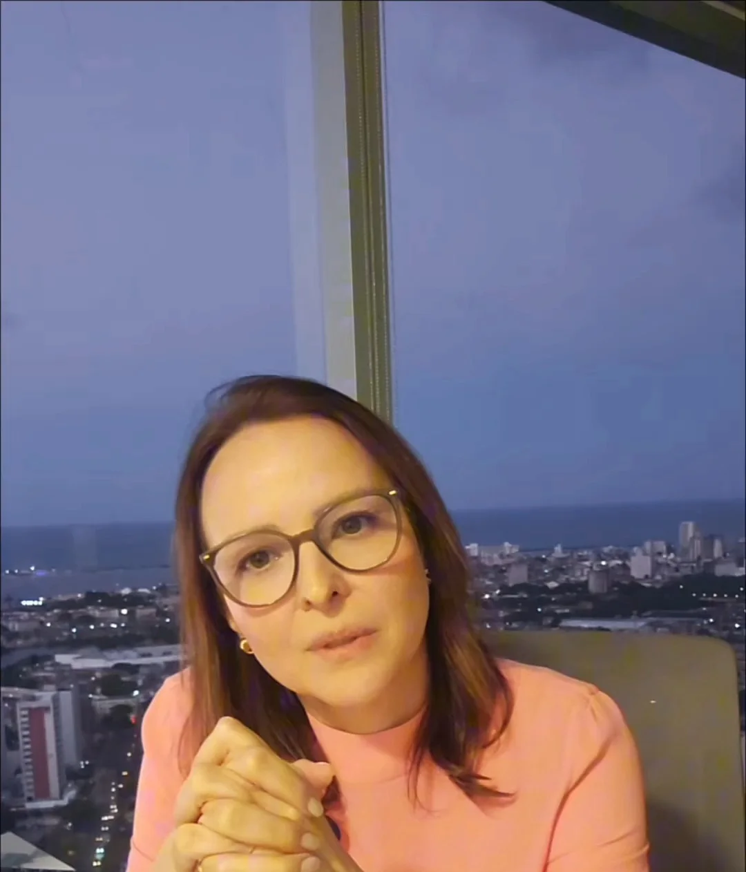 Dra. Ana Emília Pinheiro, de blusa salmão e óculos, diante de uma janela alta com a cidade do Recife e o mar ao entardecer