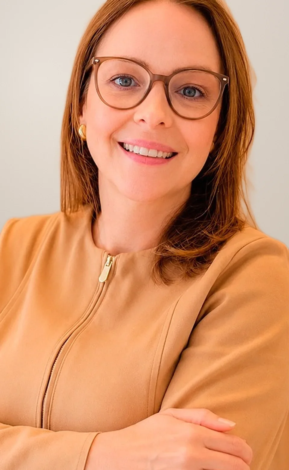 Dra. Ana Emília Pinheiro sorrindo, de óculos e blazer caramelo, com os braços cruzados